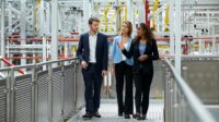 One male and two female coworkers are walking on the walkway in the factory. They are wearing business wear.