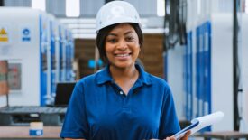 Portrait of a black female engineer technician smiling, wearing a hard hat and holding a clipboard in a warehouse packaging factory.